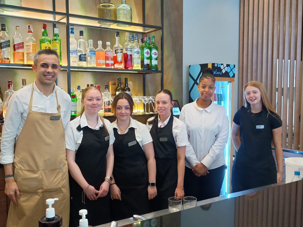 a group of students posing for a photo behind the bar at a restaurant they are working at as part of their work placement 
