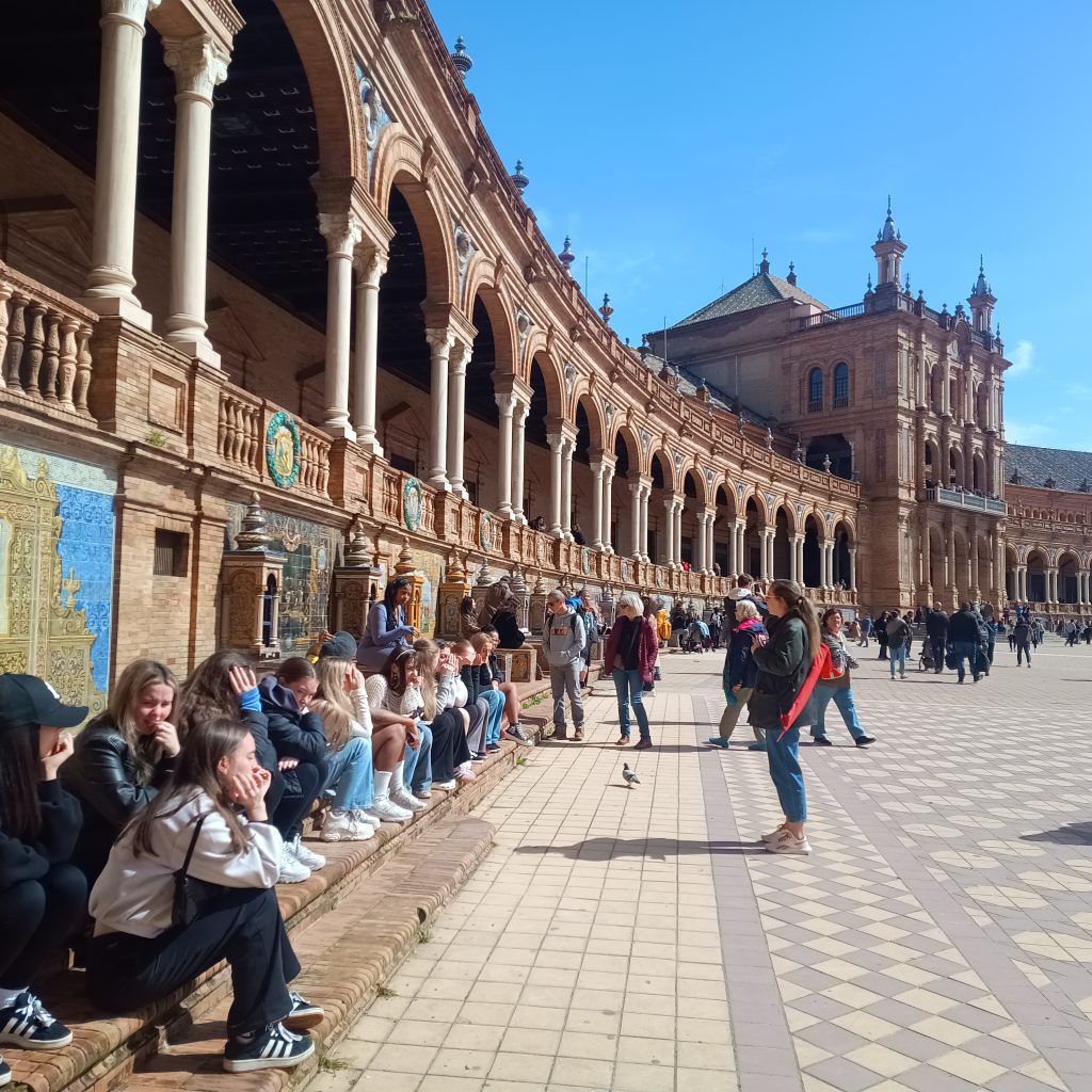 A group of students sightseeing during their trip to Seville in a large, historic plaza. There is a building with columns and arches behind. 